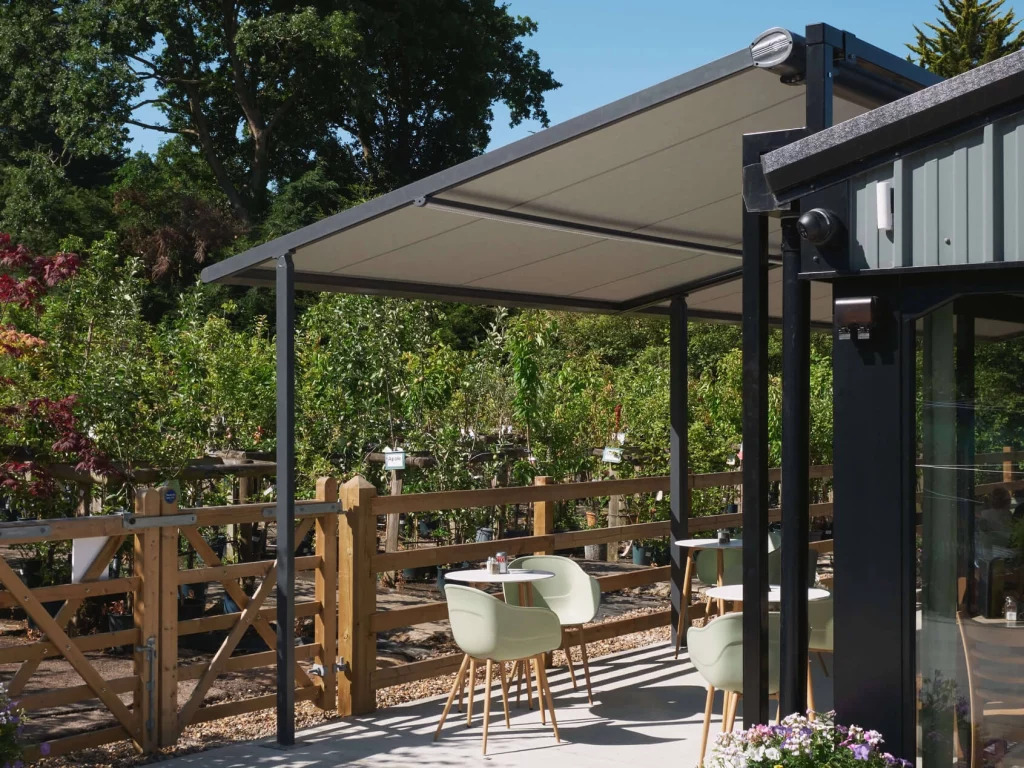Outdoor patio seating area with modern chairs under a retractable conservatory awning and wooden railing surrounded by greenery.