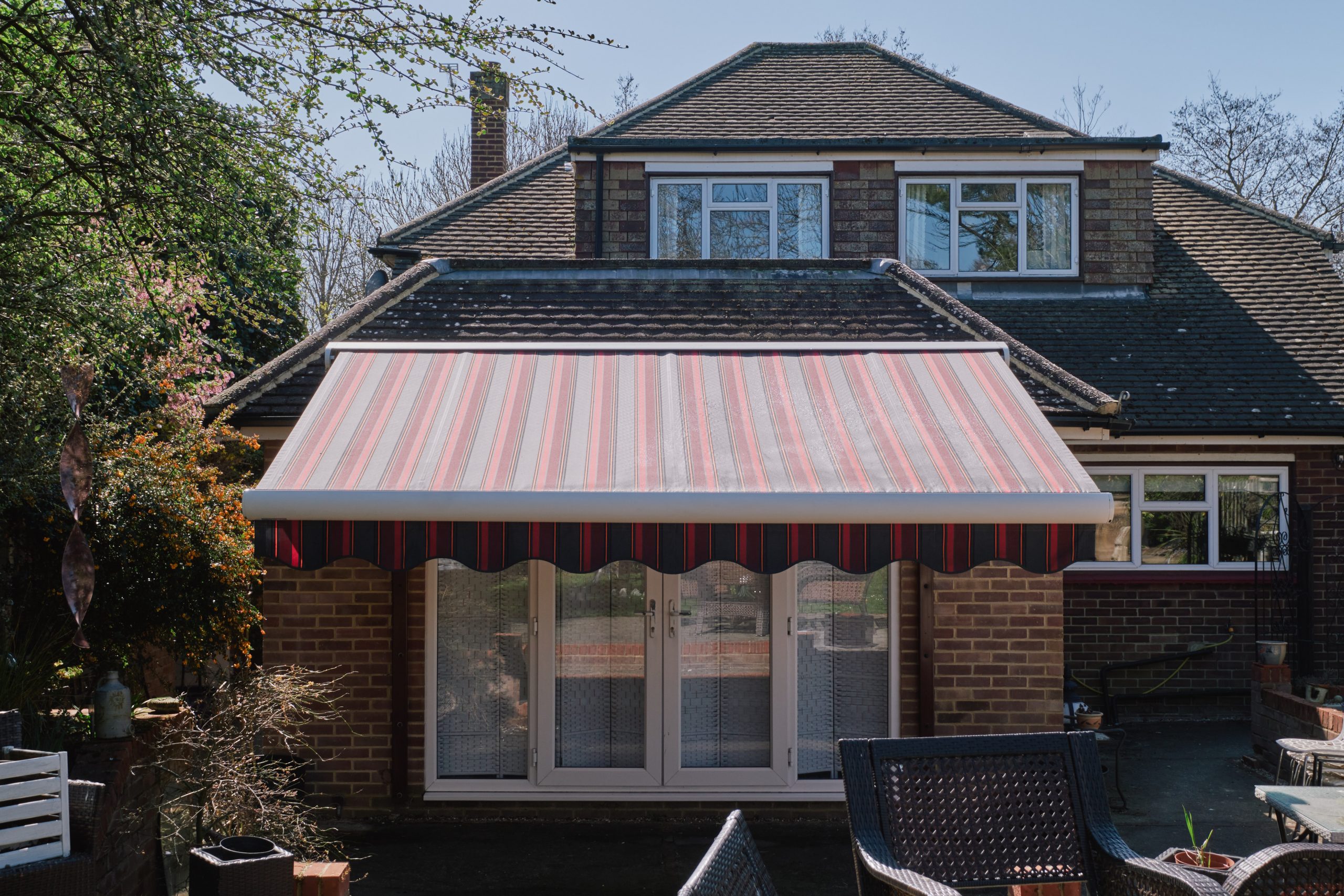 Full cassette awning with red and grey striped fabric installed above south‑facing windows, creating shaded terrace seating in a backyard.