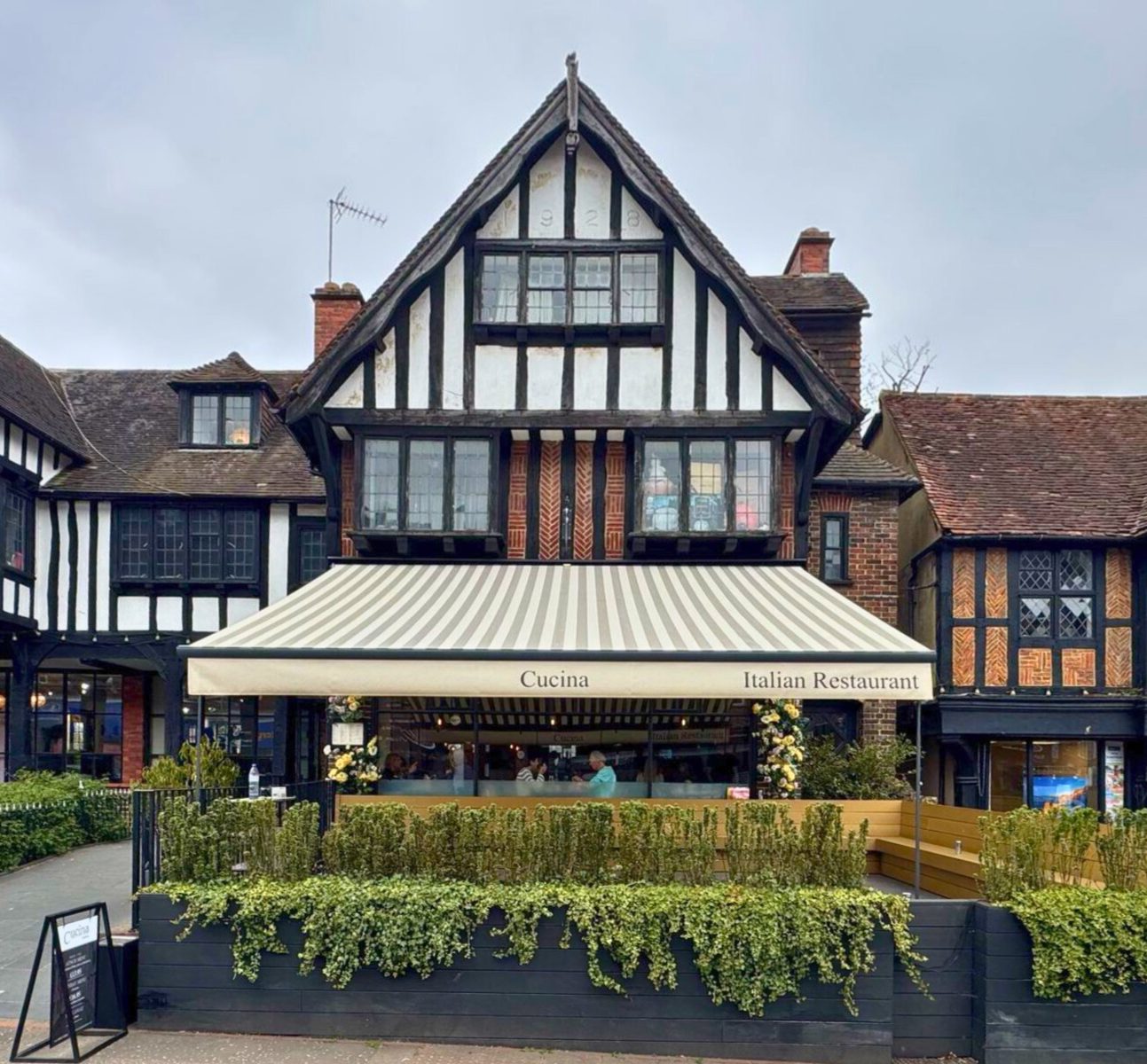 Outdoor restaurant seating area shaded by a striped beige and light brown awning. The awning has branded text on the front valance, and tables and chairs are arranged beneath it for diners.