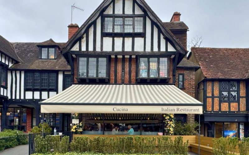 Outdoor restaurant seating area shaded by a striped beige and light brown awning. The awning has branded text on the front valance, and tables and chairs are arranged beneath it for diners.