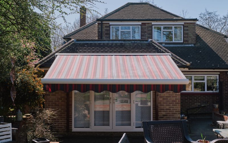 Full cassette awning with red and grey striped fabric installed above south‑facing windows, creating shaded terrace seating in a backyard.